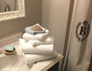 Stack of white towels and soap on the edge of a vanity sink next to a shower curtain with a B logo.