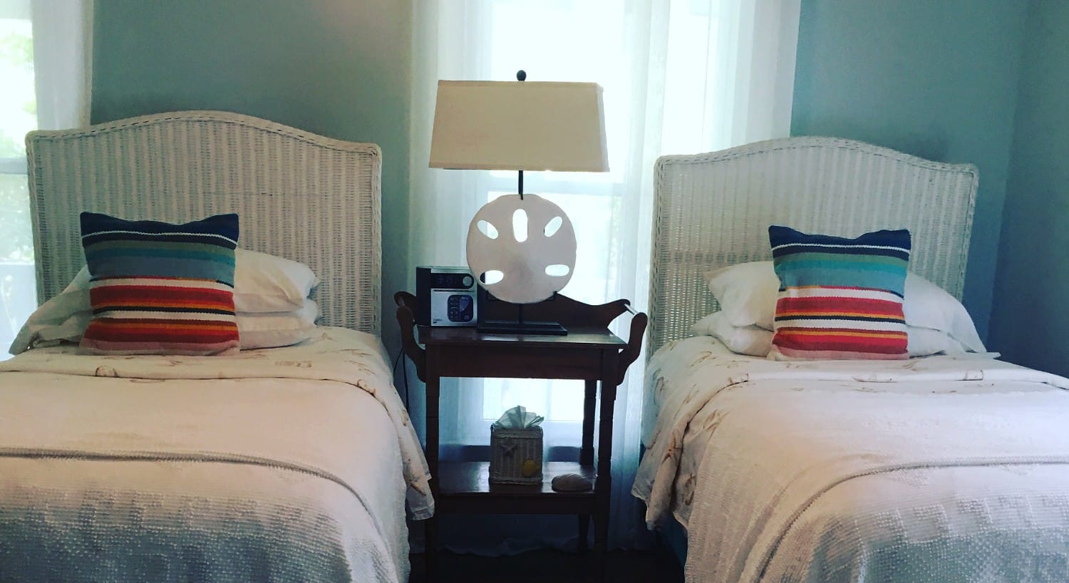 Two twin beds with white wicker headboards and white coverings with bright throw pillows. 