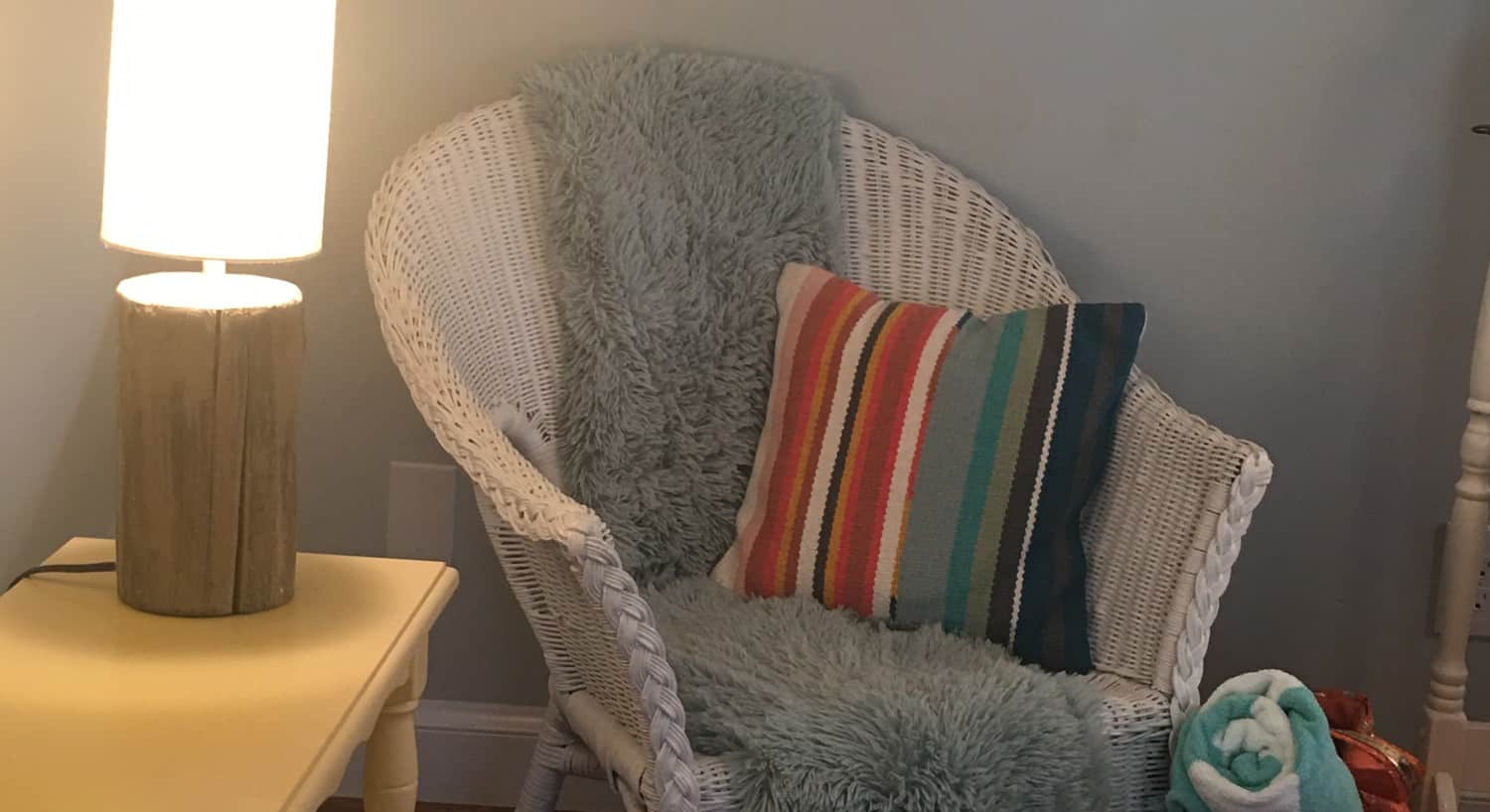 A white wicker arm chair with a cozy throw and pillow sits next to a table with a lamp. 