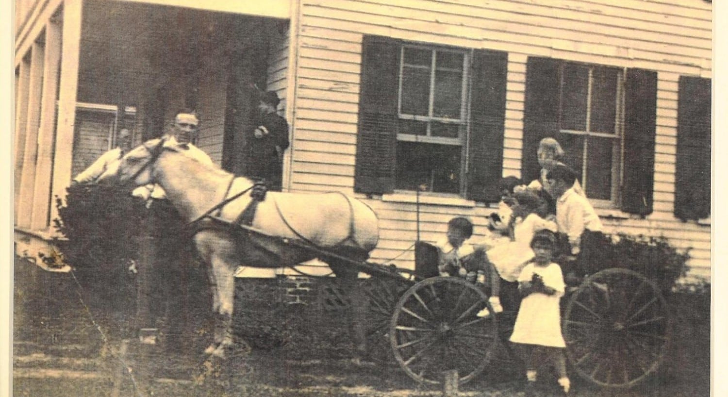 Vintage photo of a family in a horse-drawn buggy pulling up to a porch of a white house. 