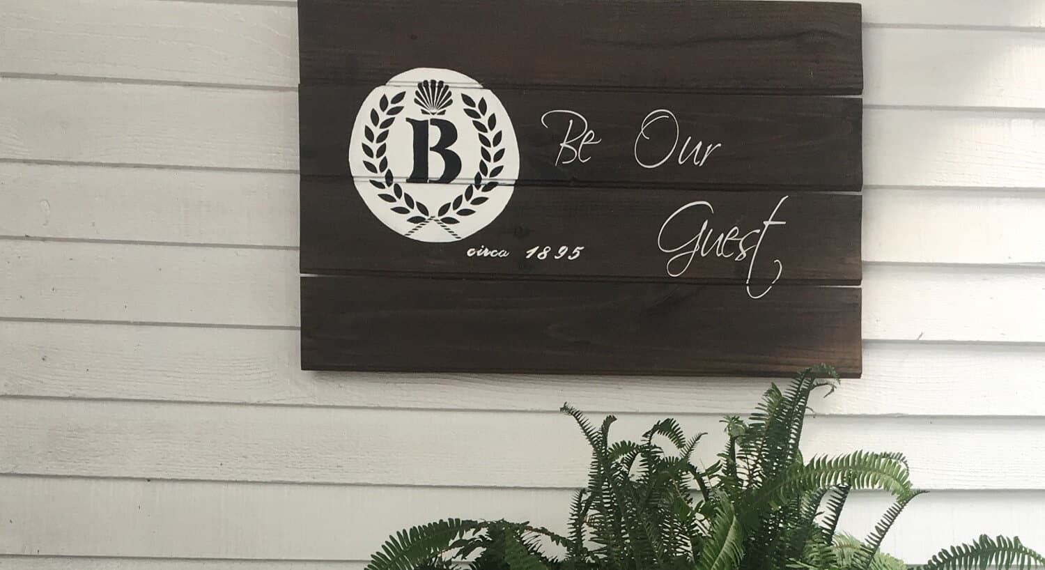 Brown wooden sign hanging on an exterior wall that says, "Be Our Guest" with the Barclay Cottage Bed & Breakfast logo. 