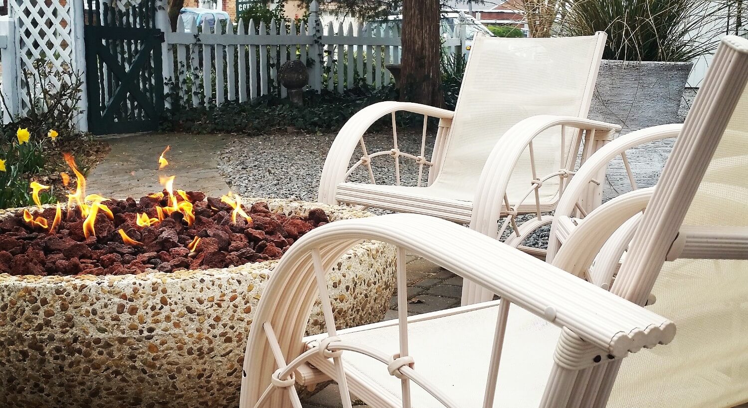 White chairs surround a unique fire pit in an outdoor patio next to a fence. 