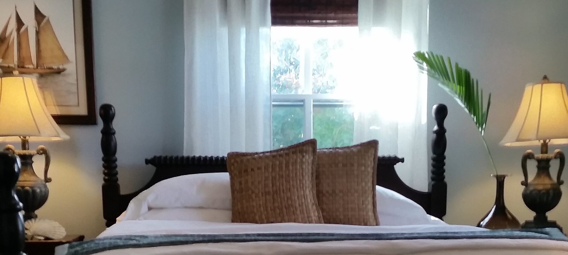 Wooden bed made up in blue and white with tan pillows between two side tables with matching lamps, under a window.