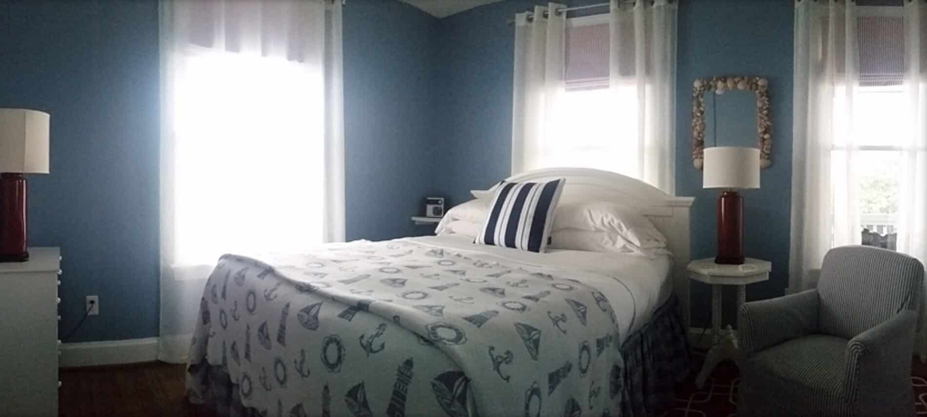 A bed made up in white and blue nautical bedding next to a nightstand with a light and an armchair.