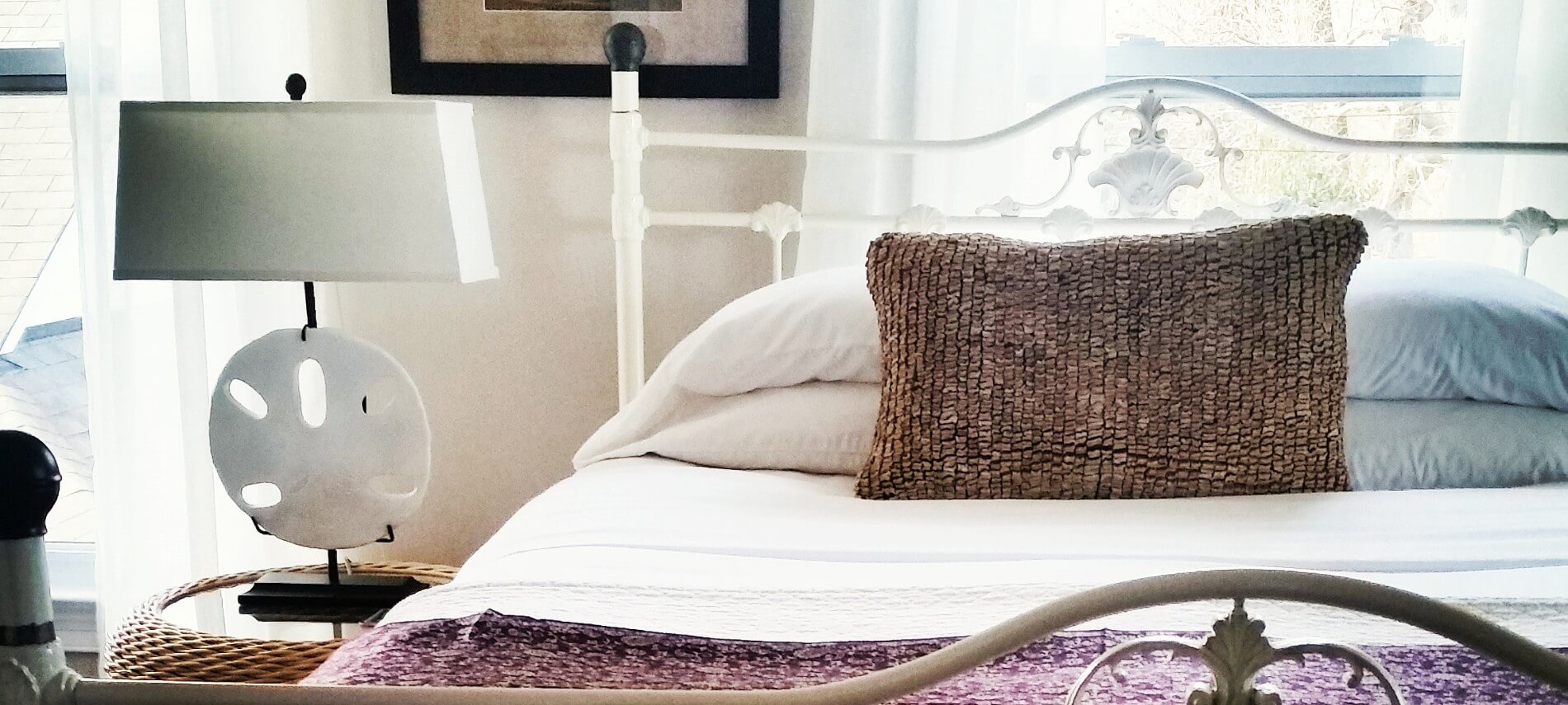 A bed with a white iron frame next to a table with a giant sand-dollar lamp.