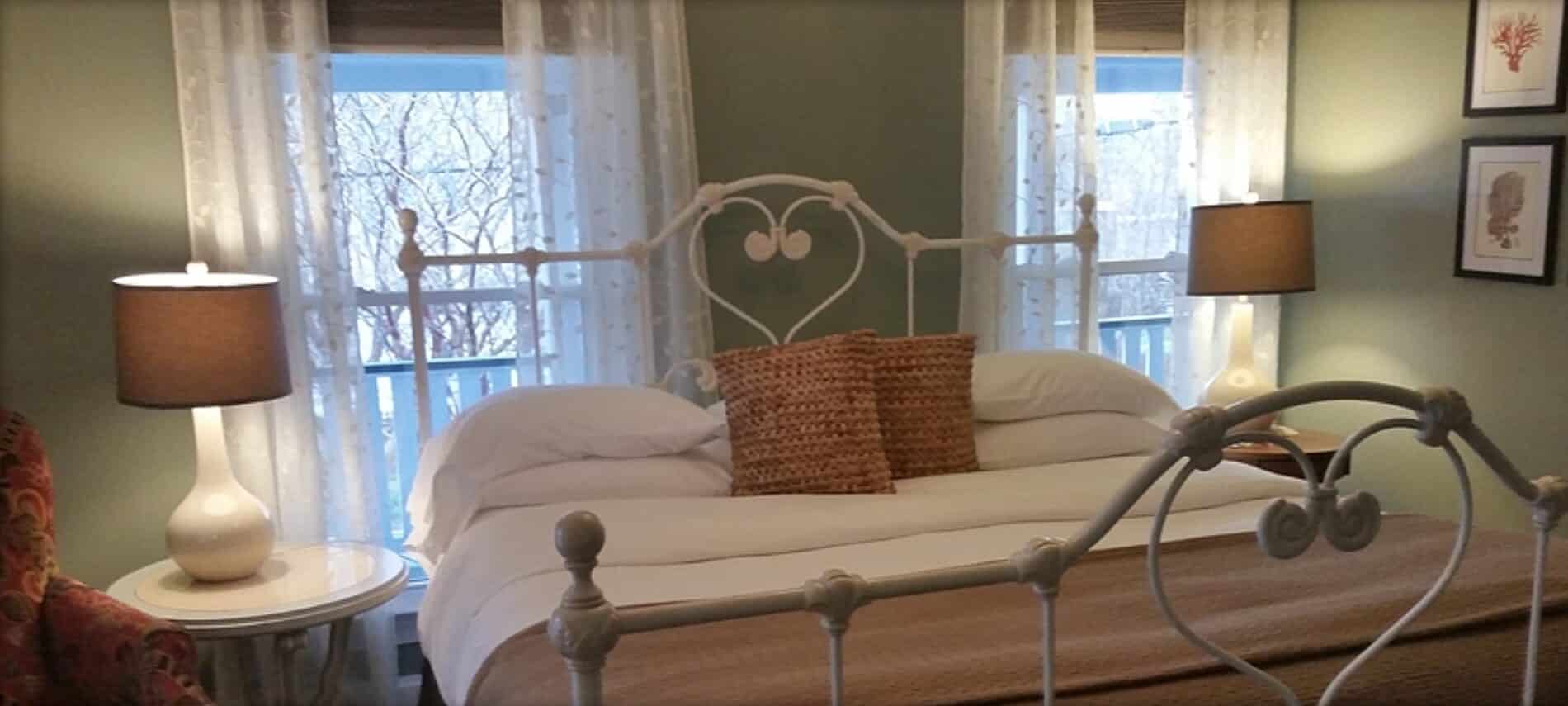 White iron bed with several pillows sits under two windows with sheer curtains.