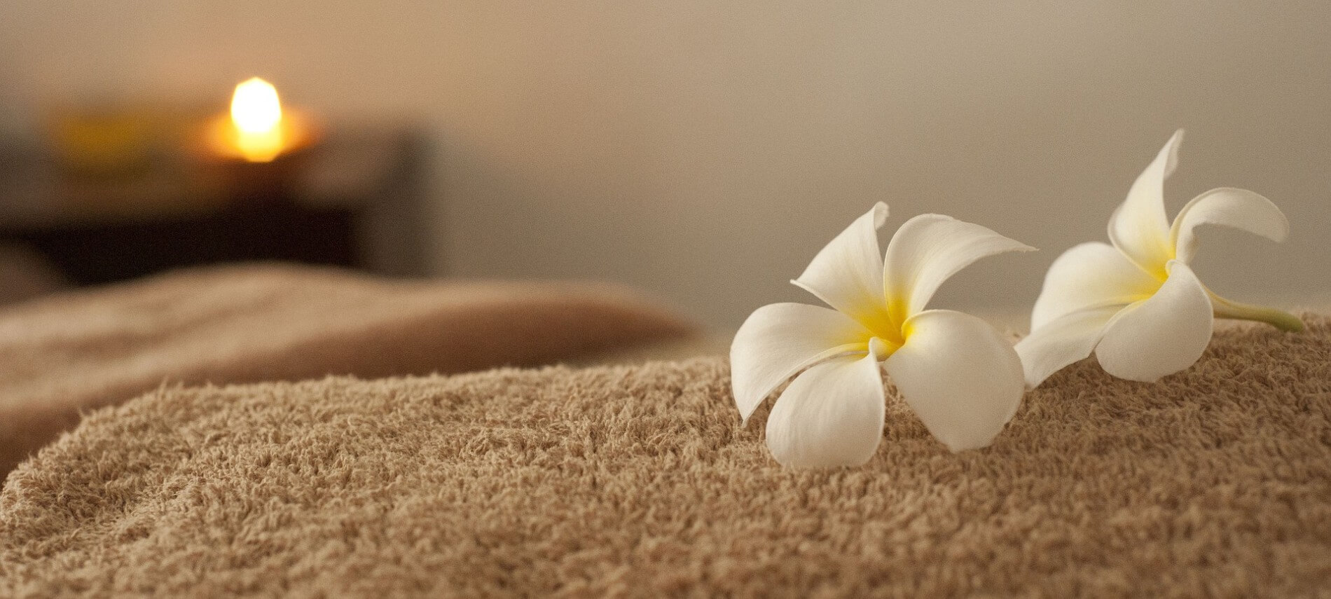 Two white flowers lie on a folded beige towel with a candle burning in the background.