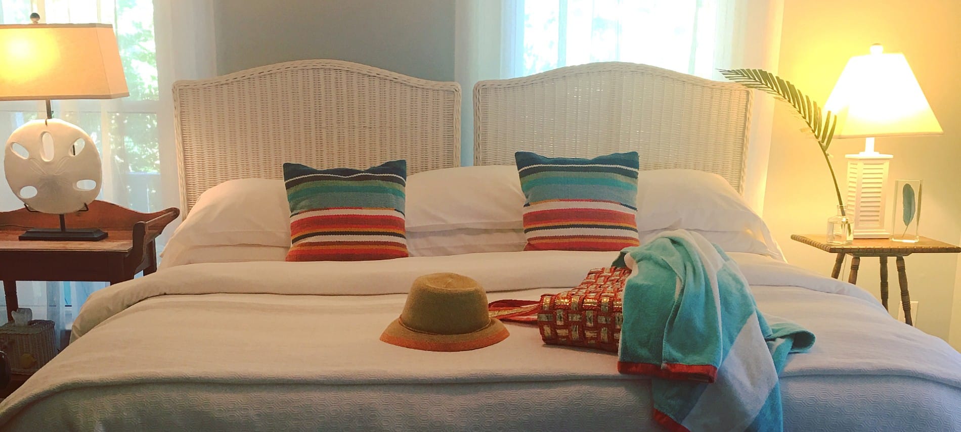 A large bed with two wicker headboards made up in white with colorful pillows and two nightstands with lamps.