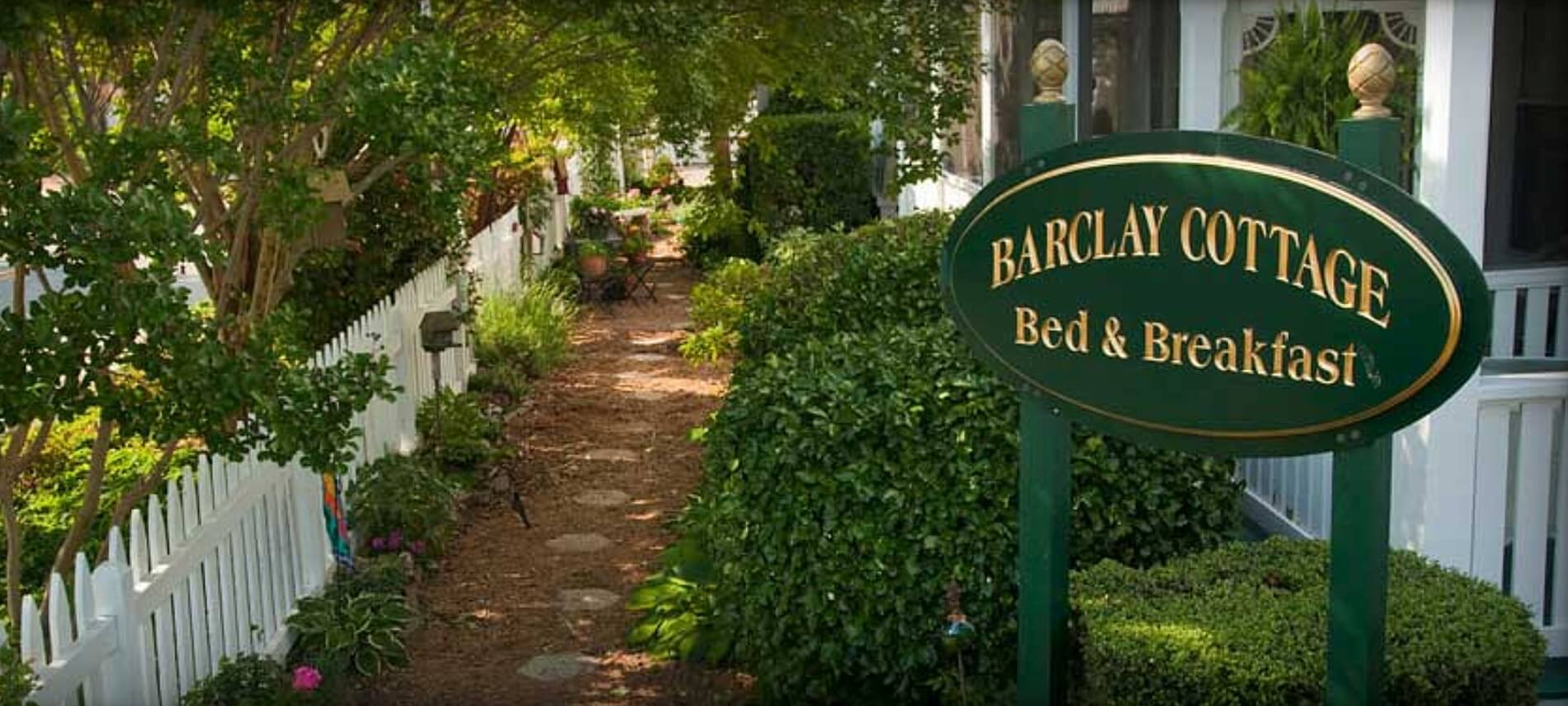 Red brick walkway between a white house and picket fence with a green and gold sign that says Barclay Cottage Bed & Breakfast 