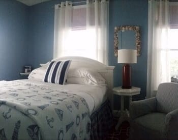 A twin bed with white and nautical bedding in a room with a side-table, armchair and two windows.