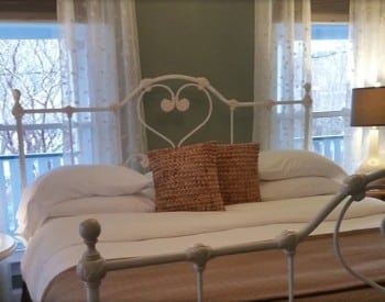 Iron bedframe with bed made up in white and tan between two large windows with sheer curtains.