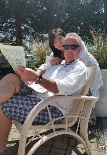 A smiling lady leans over the shoulder of a man in sunglasses reading the newspaper.