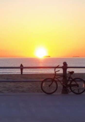 A bech cruiser rests on a fence overlooking a person on the beach at sunset.