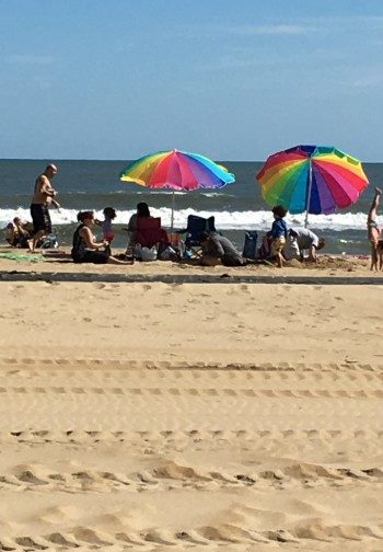 A group of people on the beach with rainbow beach umbrellas.