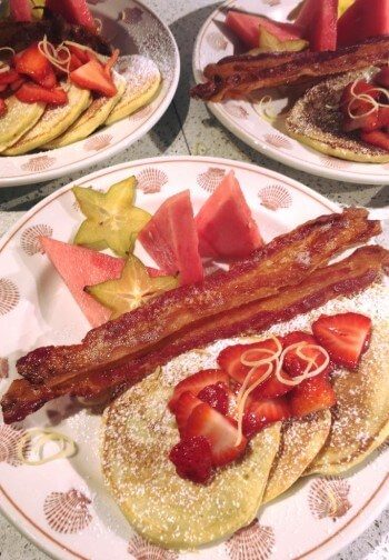 Breakfast plates with bacon, fruit and pancakes topped with cut strawberries.
