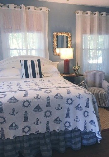 A bed made up in white and blue nautical bedding next to a nightstand with a light and a chair with striped cover.