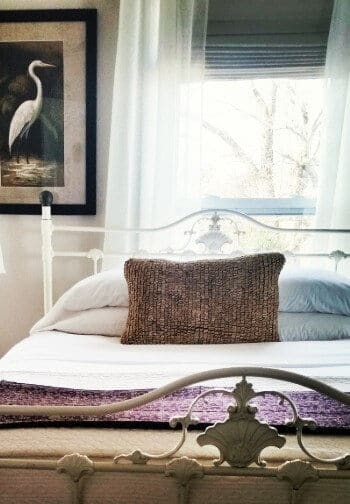 Iron bedframe with bed made up in white and tan under a large window with sheer curtains and a bird painting.