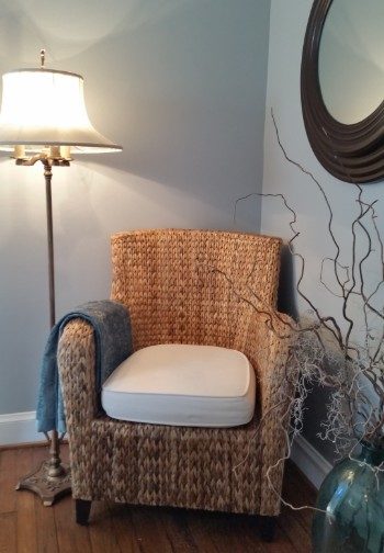 Tan wicker arm-chair with a white seat next to a stand light in the corner of the bedroom.