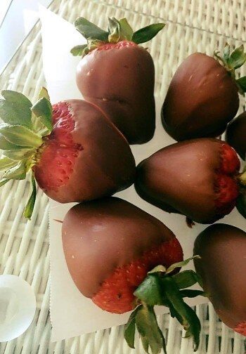 Six chocolate-covered strawberries rest on parchment over a white wicker table.