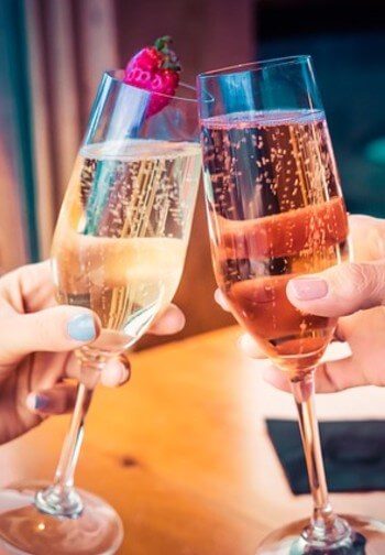 Two champagne flutes are toasted by women with pink and blue nail polish.