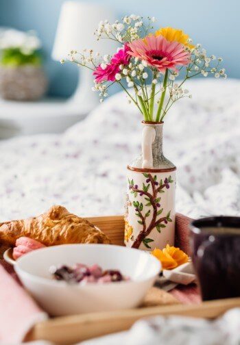 Breakfast tray with croissant and cereal alongside a cup of coffee and vase of colorful flowers.