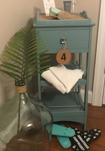 A side table painted teal next to a glass jug and two pairs of flip-flops with a #4 tag hanging from it.